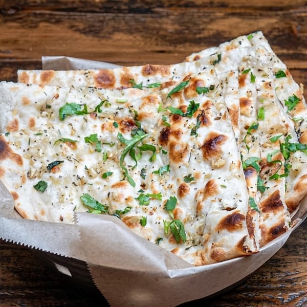 Garlic naan bread, garnished with herbs and spices, served in a paper-lined basket on a wooden table.