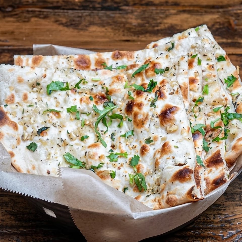 Garlic naan bread, garnished with herbs and spices, served in a paper-lined basket on a wooden table.